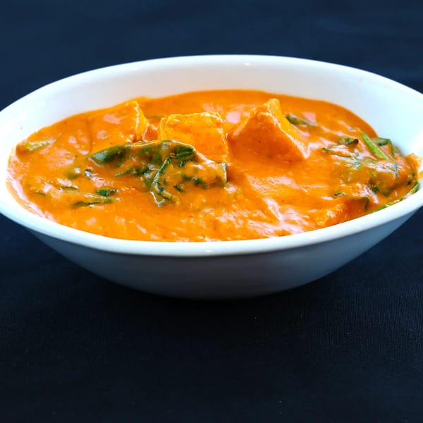 A white bowl filled with vibrant orange Indian curry, featuring paneer and spinach, on a dark background.